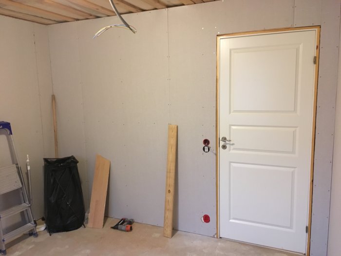 Renoverat rum med nygipsade väggar, uttag för takdosan, stängd dörr och renoveringsmaterial på golvet.