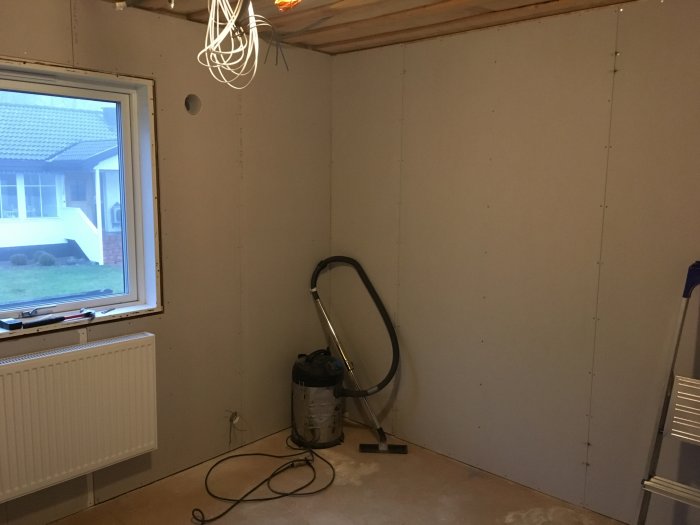 Gipsvägg i ett rum under renovering med synliga skruvar, ledningar för el och en dammsugare i hörnet.