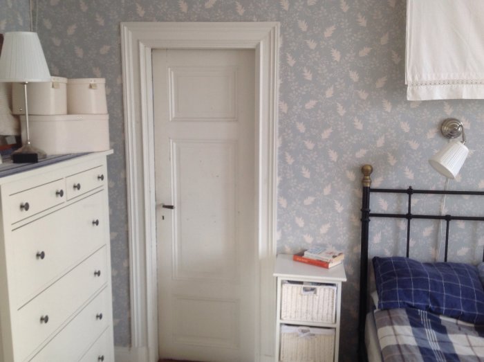 Sovrum med originaldörr från 1900-talet, vit byrå, metall sängram och blå-vit tapet.