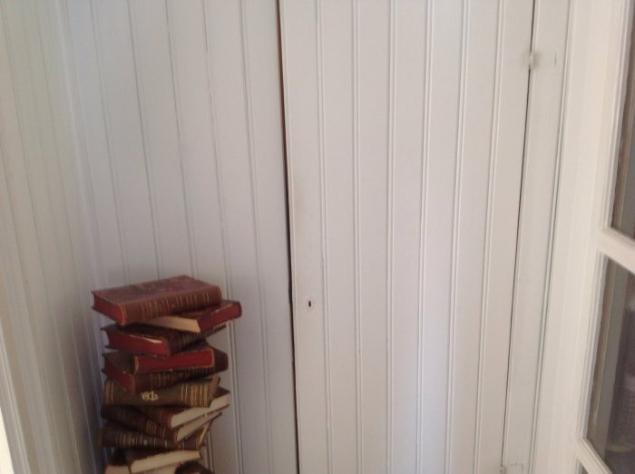 Stängd dörr till linneskåp bredvid en stapel gamla böcker, vit vägg och fönsterkarm.