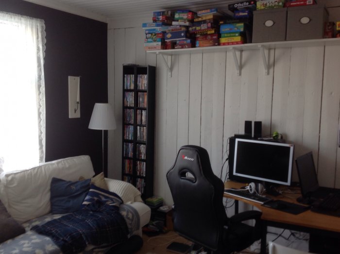 Lekrum med vitt målade plankväggar, hyllor med spel, soffa, gamingstol och skrivbord med dator.