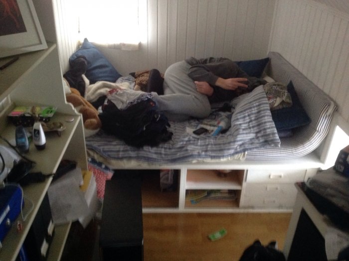 Ostädat tonårsrum med kläder och föremål utspridda, person ligger på en bäddsoffa.