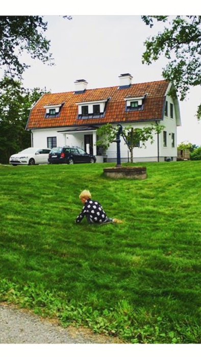 Vit villa med tegeltak, barn i förgrunden på en gräsmatta, bil parkerad intill huset.