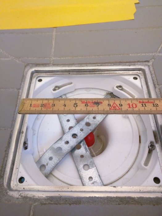 Mätning av en golvbrunnens diameter med en tumstock, bredvid ett nuvarande luktstopp.