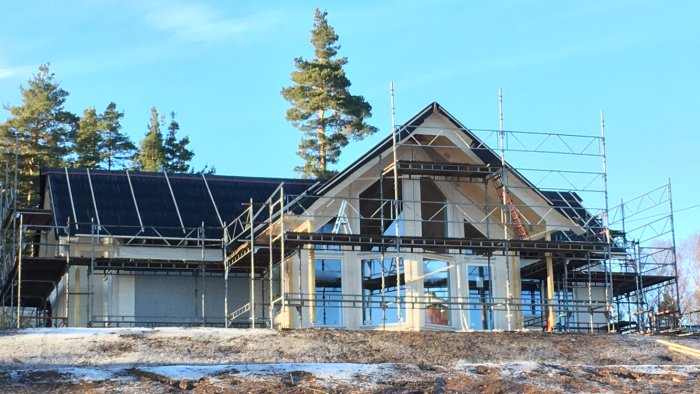 Hus under konstruktion med nylagt takpapp, omgivet av byggnadsställningar mot skogsbakgrund.