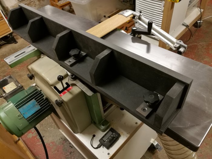 Anhåll monterat i utmatningsbordet på snickerimaskin med stålspännvred och svarta skyddande komponenter.