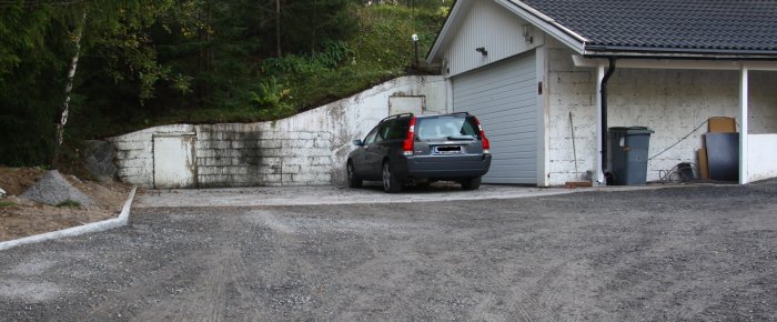 Bild av en uppfart med en parkerad bil framför en sliten vit betongvägg med lutning, bredvid en garagebyggnad.