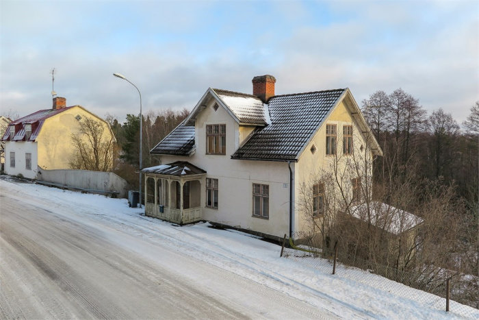 Äldre tvåvåningshus med snötäckt tak och veranda, belägen vid en snöig väg omgiven av träd.