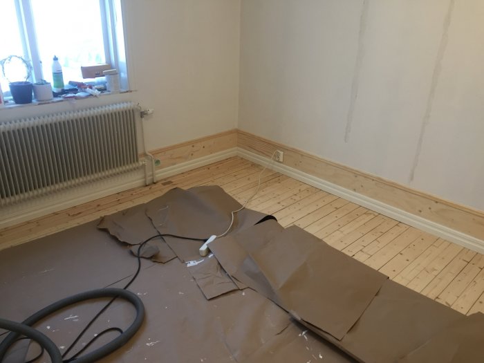 Renoveringsrum med ommålad vägg, trägolv, radiator och golvsocklar under renovering.