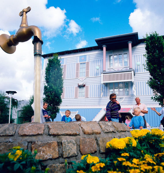 Ett upp och nervänt hus vid Liseberg med en svävande kran som porlar vatten, omgivet av besökare och blomster.