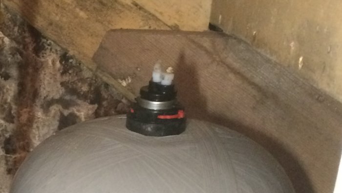 Topp av en hydrofor med inbyggd flottör och två plastskruvar, en med nippel, mot en bakgrund av isolering.