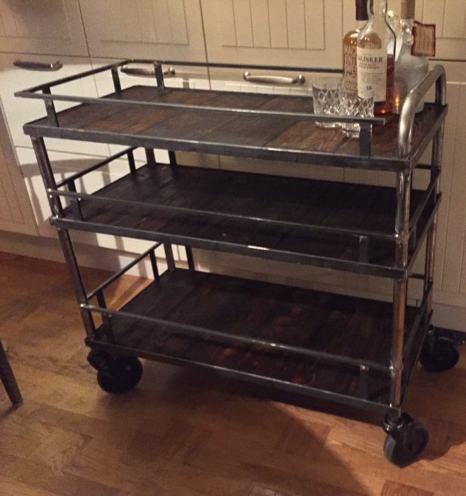 Nybyggd drinkvagn i metall med trähyllor på hjul, whisky och glas ovanpå.