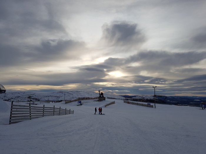 Vinterlandskap med moln över Vemdalsskalet, solnedgång och skidåkare på snötäckt backe.