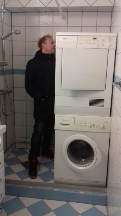 Man i badrum med staplad tvättmaskin och torktumlare framför kakelklädd vägg.