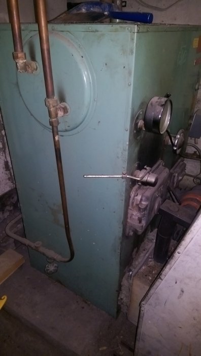Äldre värmepanna i metall med synliga röranslutningar och övergivet utseende står i ett grovkök eller pannrum.