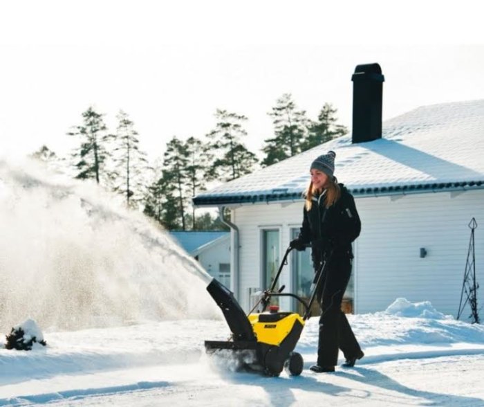 Person använder gul och svart Stiga snöslunga på snötäckt uppfart med hus i bakgrunden.