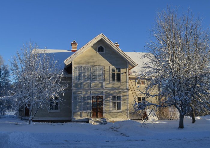 Tvåvånings trävilla täckt av snö med barträd i förgrunden och blå himmel ovanför, i begynnande renoveringsprojekt.