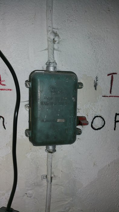 Äldre grön elektrisk dosa på en vägg med anslutna metallrör och elledningar, tecken på åldring och användning.