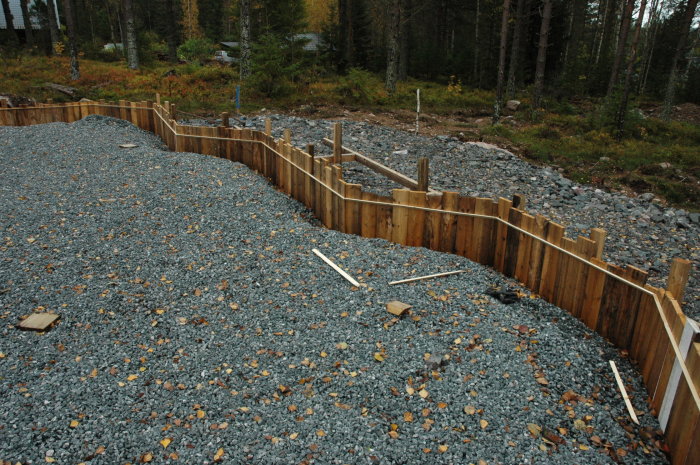 Byggplats med grus och träformar för gjutning av fundament i skogsmiljö.