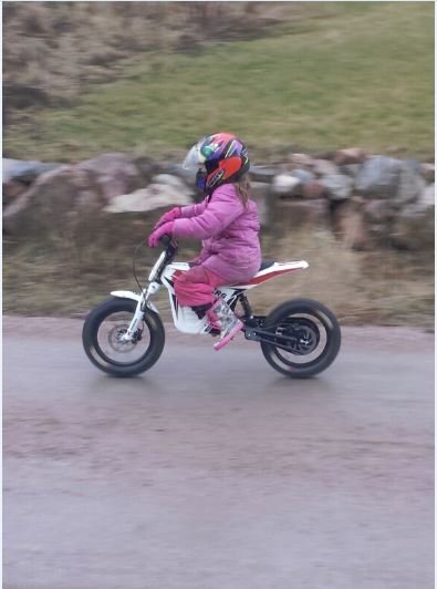 Barn i rosa ytterkläder kör minicrossmotorcykel på en väg med oskarp bakgrund.