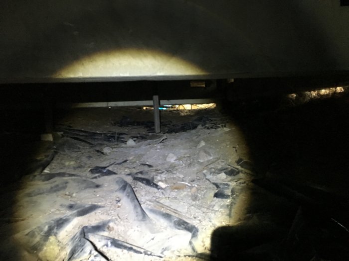 Krypgrund med slarvigt gjutna plintar, brädor, trasig plast och avfall synligt under huset.