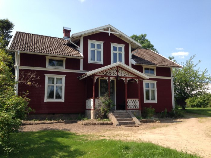 Rödmålat hus från tidigt 1900-tal med vita knutar och dekorerad veranda i lantlig miljö.
