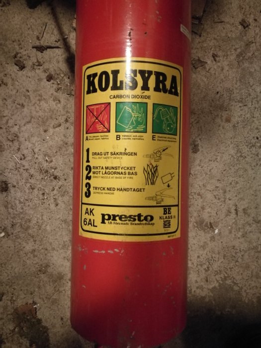 Röd kolsyrebehållare med varningsetiketter och texten 'KOLSYRA' på en grov betongyta.