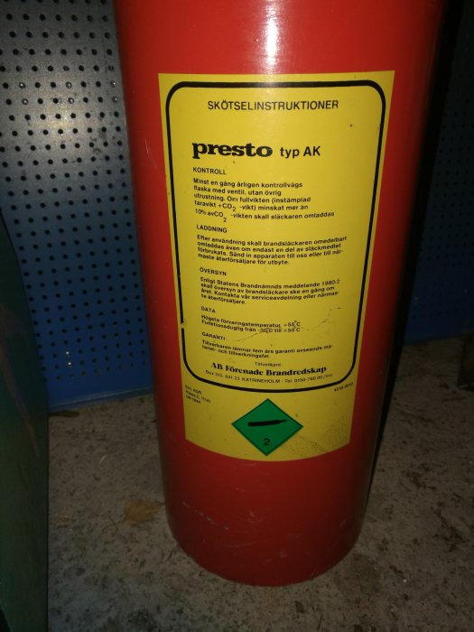 Röd kolsyretub av typen Presto AK med skötselinstruktioner och brandklassningsetiketter i ett garage.