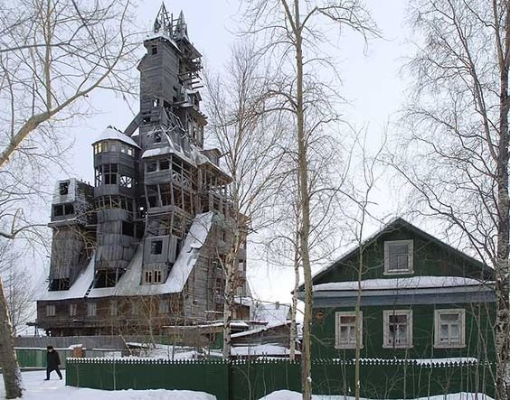 En excentrisk och komplex trästruktur bakom ett traditionellt grönt hus omgivet av snö och träd.