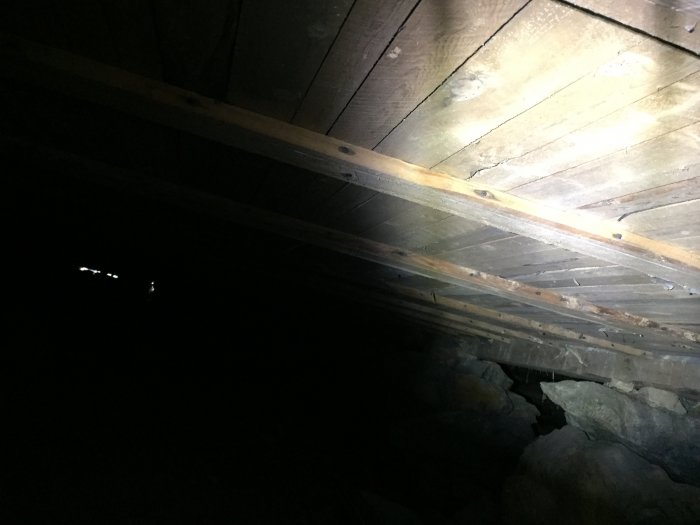 Mörk krypgrund under hus med synliga träbjälkar och stenar, potentiellt för isolering.