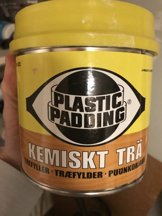 Burk med texten "PLASTIC PADDING KEMISKT TRÄ" hållen i handen.