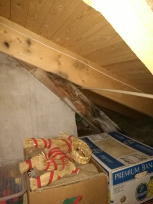 Mögliga takstolar på en vind med synliga svarta fläckar bredvid nyare ljust virke, omgiven av lagrade föremål.