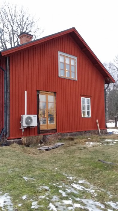 Rödmålat trähus med vita fönster och en värmepump på fasaden, delvis snötäckt gräsmatta i förgrunden.