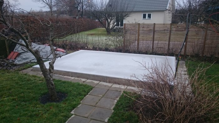 Nyggjuten isolerad betongplatta för förråd i trädgård, omgiven av gräsmatta och staket.