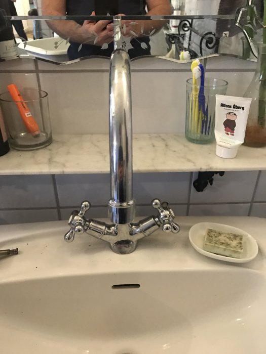 Tvättställsblandare med tappat strålsamlare framför en spegel på ett badrum, med tandborstar och tandkräm på hyllan.