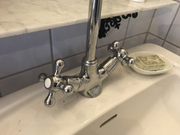 Tvättställsblandare med korsformade handtag och skadad strålsamlare på ett handfat.