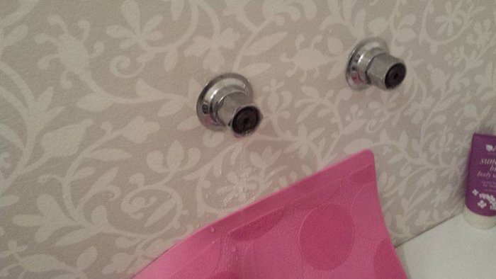 Två vattenkranar utan filter i en kakelvägg ovanför en del av en rosa duschdraperi.