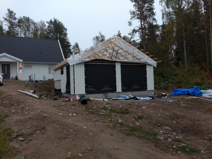 Byggplats med halvfärdig byggnad där takstolar är synliga, omgiven av skog och närliggande hus.