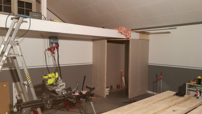 Inomhus byggplats med gipsskivor och verktyg framför en oavslutad väggsektion.