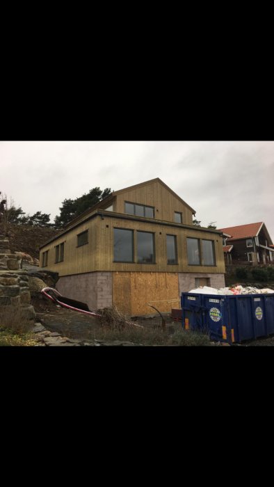 Nybyggt hus med stort garage under konstruktion på en smal tomt, med sopcontainrar framför.