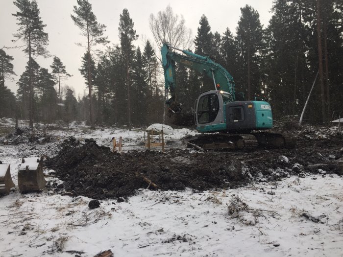 Grävmaskin på arbetsplats med snötäckt mark, förbereder för gjutning av platta och markarbete under carport.