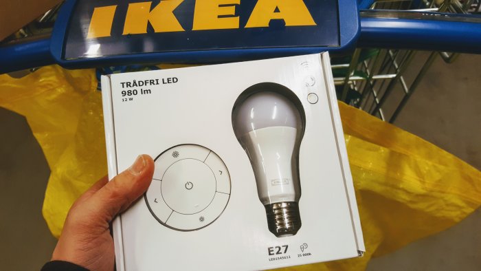 Hand håller en förpackning av IKEA Trådfri LED-lampa med fjärrkontroll vid en kundvagn och IKEA-varukasse.