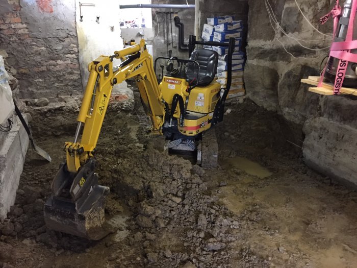 Minigrävmaskin i arbete med urgrävning av källargolv, omgiven av lerig jord och delvis gjutna stödmurar.