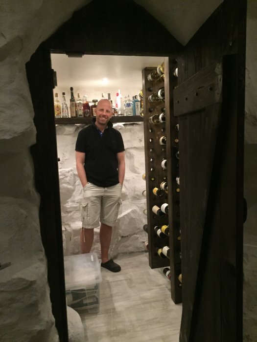 Renoverad källare med vitmålade naturstensväggar och nytt golv, man framför vinställ.