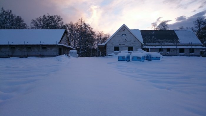 Byggfas av ett garage i vinterskymning med snötäckt tak och omgivning, synliga takstolar, ej pålagda takpannor.