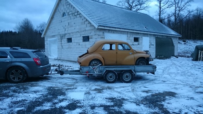 En gul klassisk bil på en släpvagn framför ett oputsat garage i två våningar med snötäckt mark runtomkring.