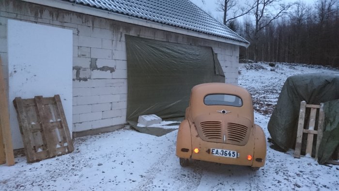 Klassisk bil parkerad framför ett halvfärdigt garage med snötäckt mark och skog i bakgrunden.