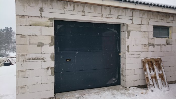 Nyinstallerad garageport i mörkgrå, på en vägg under konstruktion, under lätt snöfall.