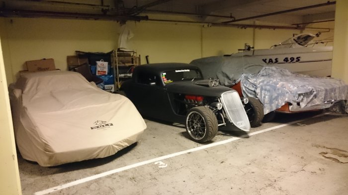 Hot rod utan täckduk parkerad i ett garage bredvid bilar och en båt under presenningar.