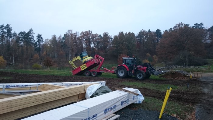 Traktor och dumprar i arbete med att flytta jord på lerig byggarbetsplats, omgivna av träd.